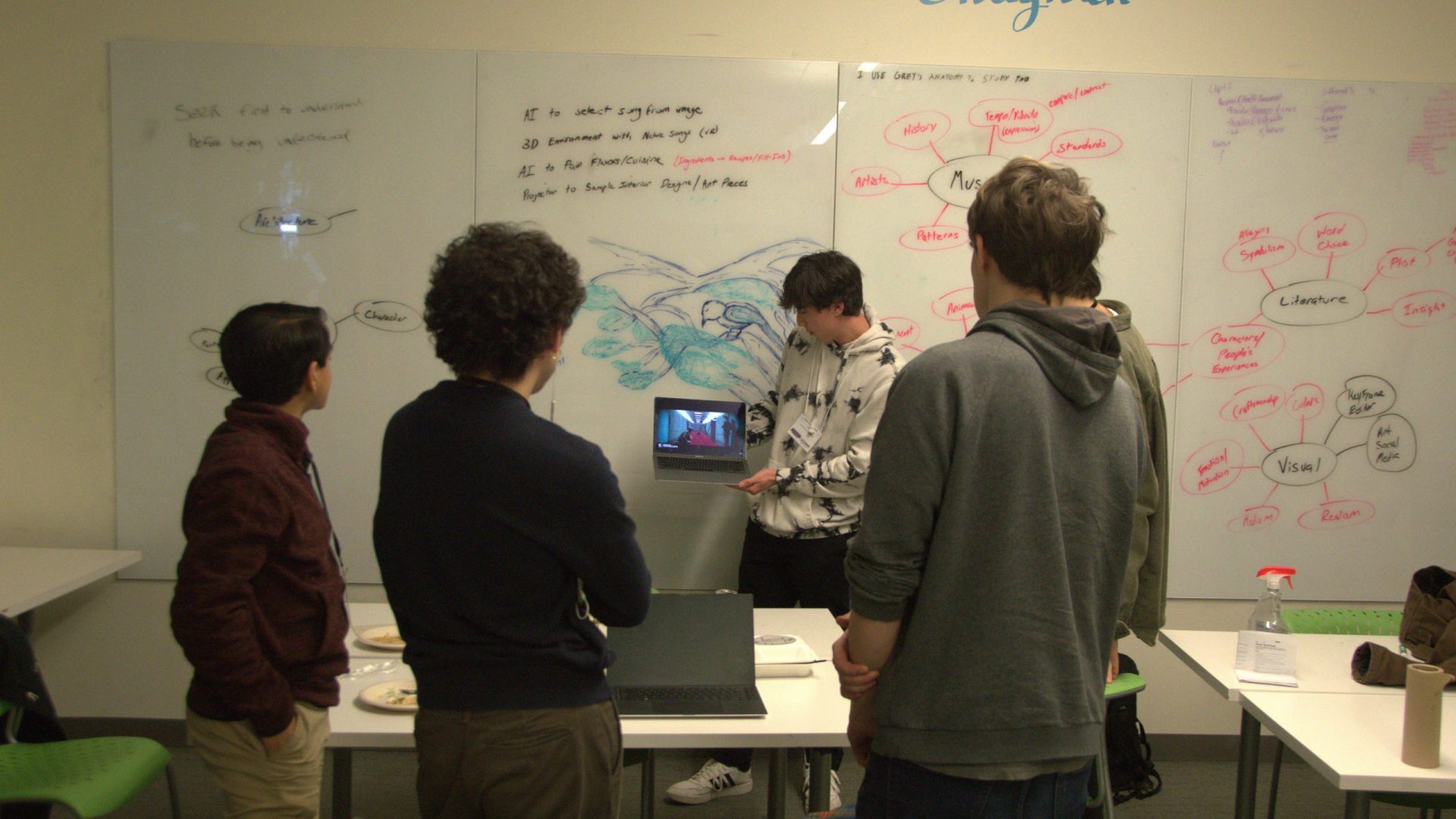 A group of studnets stand around a desk with a student holding up a laptop as they begin planning their innovation